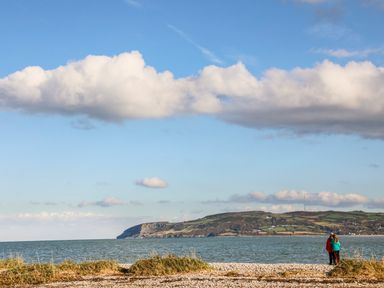 Two people standing on a pebbled beach near grass with water and hills in the background at Neaudd Wen in Red Wharf Bay near Benllech