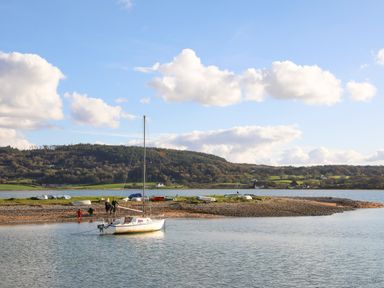 A small sailboat anchored near a rocky shore with people standing on the shore and hills in the background at Neaudd Wen Red Wharf Bay near Benllech