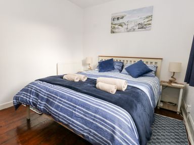 A bedroom with a double bed striped blue bedding two beige rolled towels two blue pillows two bedside tables with lamps and a beach painting on the wall at Saddlers Cottage in Weymouth