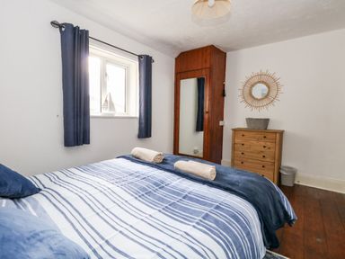 A bedroom with a bed striped blue and white bedding a wooden dresser a round mirror and a window with blue curtains at Saddlers Cottage in Weymouth