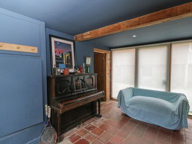 A room with a black upright piano decorated with bottles and a framed picture and a blue covered sofa near large windows at Brandeers Long Barn in Brandier near Minety