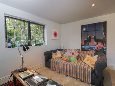 A living room with a sofa covered with a checkered throw and cushions with a wooden table and books and a lamp near a window with glass bottles and artwork on the walls at Brandeers Long Barn Brandier near Minety