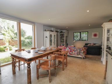 A dining area with a wooden table and chairs next to a living room with sofas and a bookshelf at Brandeers Long Barn in Brandier near Minety