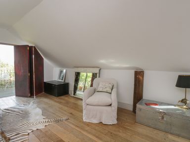 An attic room with a white armchair a black cabinet a large mirror a metal chest and a table lamp at Brandeers Long Barn in Brandier near Minety