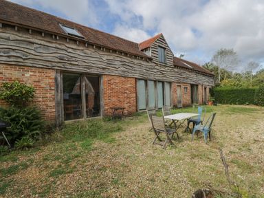 An outdoor area with a table and mixed chairs in front of a long brick and wooden barn building at Brandeers Long Barn Brandier near Minety