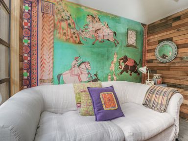 A room with a white sofa with colorful pillows a wooden wall a wall painting showing people on horses and an elephant and a round mirror at The Big Box Brandeers Brandier near Minety