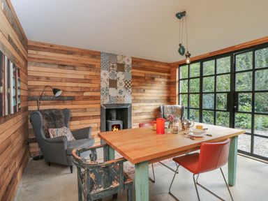 A dining room with a wooden table, mixed chairs, wood-paneled walls, a fireplace, and large windows showing greenery at The Big Box Brandeers in Brandier near Minety