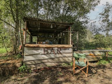 An outdoor wooden bar structure surrounded by trees with a green chair in front at The Big Box Brandeers Brandier near Minety