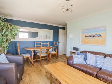 A living room with a brown leather sofa with blue striped cushions a wooden coffee table a wooden dining table with chairs a tall floor lamp and a large mirror on a blue wall at Seaspray in St Bees