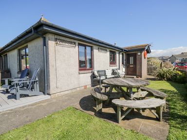 An outdoor patio area with wooden picnic table and benches and two grey chairs outside a single-story house at Seaspray in St Bees