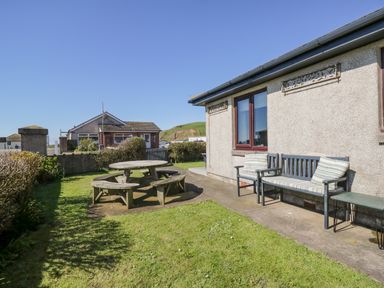 An outdoor garden area with wooden round picnic table and benches and a patio with cushioned chairs at Seaspray in St Bees