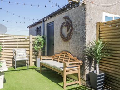 A small outdoor patio with a wooden bench and chair with cushions artificial grass potted plants string lights on a wall at Seaspray in St Bees