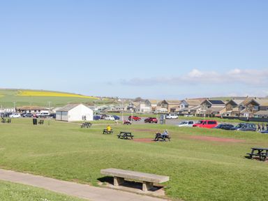 A park with benches and picnic tables and houses and cars in the background at Seaspray in St Bees