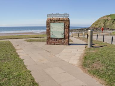 A path leading to a stone information board near the sea with a grassy hill in the background at Seaspray in St Bees