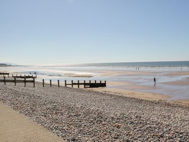 A pebble beach with wooden groynes leading to a sandy shore and people walking near the water at Seaspray in St Bees