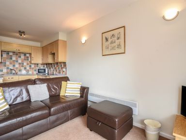 A living room with a brown leather couch, striped cushions, a brown ottoman, a small kitchen with tiled backsplash, and a framed picture on the wall at 3 Bay View Court in Lyme Regis