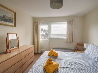 A bedroom with a bed covered in chevron patterned bedding and yellow towels on top a wooden dresser with a mirror a chair and a window with curtains overlooking the sea at 3 Bay View Court in Lyme Regis