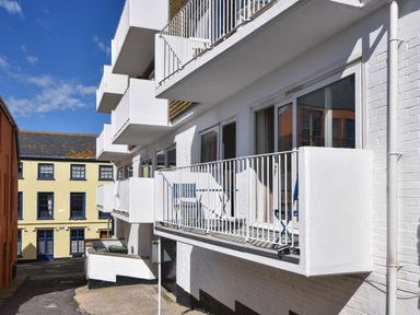 A row of white balconies with railings attached to a white brick building next to a yellow building with blue window frames at 3 Bay View Court in Lyme Regis