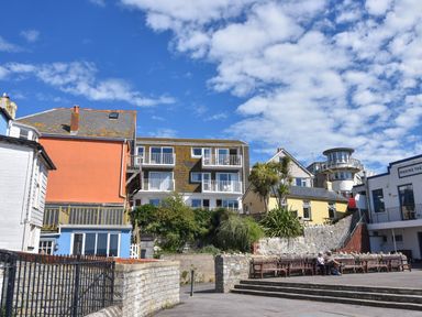 A group of buildings with balconies and stairs by a paved area with benches and people sitting at 3 Bay View Court Lyme Regis
