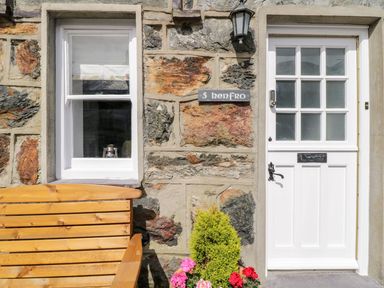 A stone wall with a white door and window a wooden bench and flowers at Henfro in Beddgelert