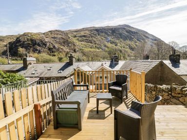 An outdoor wooden deck with chairs a bench and a small table overlooking rooftops and a hillside at Henfro in Beddgelert