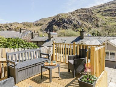 An outdoor deck with a bench chair table and flowers overlooking rooftops and hills at Henfro in Beddgelert