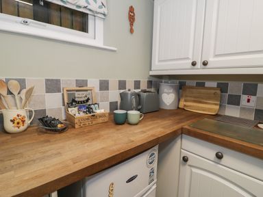 A kitchen countertop with wooden utensils in a jug tea and coffee sachets in a wooden storage box two mugs a kettle a toaster a bread container and cutting boards at Henfro in Beddgelert