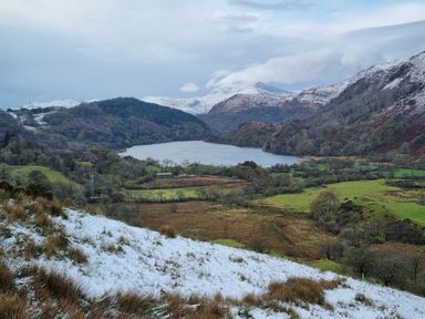 A lake surrounded by trees and hills with some snow on the ground and mountains in the background at Henfro in Beddgelert