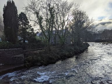 A river flowing beside leafless trees and houses with hills in the background at Henfro in Beddgelert