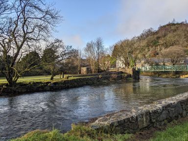 A river with stone walls along the bank trees and a metal footbridge over the water at Henfro in Beddgelert