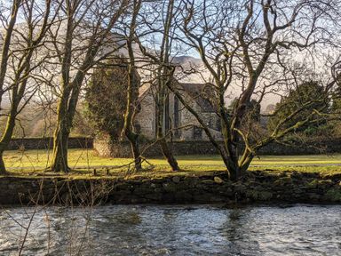 A stone building behind leafless trees near a river with a stone wall in Henfro Beddgelert