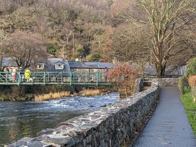 A river with a stone wall and pathway beside it and a metal pedestrian bridge with people crossing at Henfro in Beddgelert