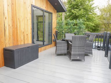 An outdoor deck with grey wicker chairs around a table and a black storage box at Watch Tree Lodge in Allithwaite near Cartmel