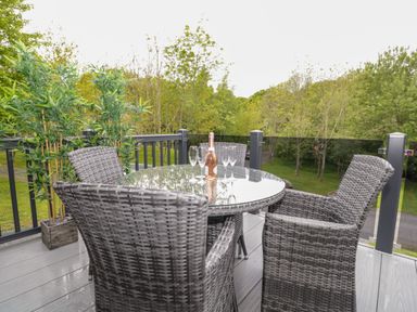 A balcony with a round glass table surrounded by four woven chairs and a bottle with glasses near plants at Watch Tree Lodge in Allithwaite near Cartmel