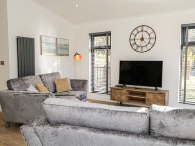 A living room with grey sofas a wooden TV stand a wall clock and two windows at Watch Tree Lodge in Allithwaite near Cartmel