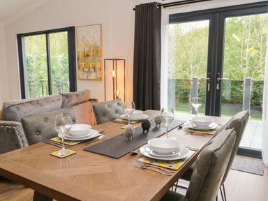 A dining table set with plates bowls wine glasses and cutlery in a room with large windows and a sofa at Watch Tree Lodge in Allithwaite near Cartmel