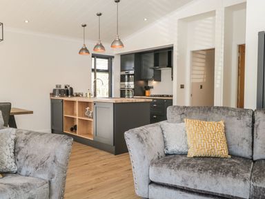A living room with grey sofas and pillows viewed towards a kitchen with an island and pendant lights at Watch Tree Lodge in Allithwaite near Cartmel