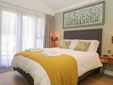 A bedroom with a bed covered in white and yellow blankets two cushions a wooden bedside table and a large window with sheer curtains at Watch Tree Lodge Allithwaite near Cartmel