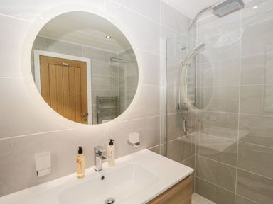 A bathroom with a round mirror above a sink and a shower area with a glass screen at Watch Tree Lodge in Allithwaite near Cartmel