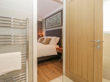 A view from a bathroom showing a wooden door open to a bedroom with a bed and artwork above it at Watch Tree Lodge in Allithwaite near Cartmel