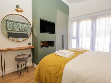 A bedroom with a bed covered in white and yellow blankets towels on the bed a wall-mounted tv fireplace round mirror wooden stool and sheer curtains at Watch Tree Lodge in Allithwaite near Cartmel