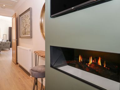 A hallway with wooden floor a wall-mounted electric fireplace and television a grey stool wooden table and framed artwork at Watch Tree Lodge Allithwaite near Cartmel