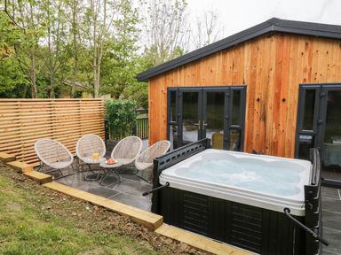 An outdoor patio with a hot tub four chairs and a wooden cabin at Watch Tree Lodge in Allithwaite near Cartmel