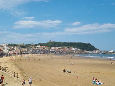 A sandy beach with people walking and relaxing with a town and a hill with trees in the background at The Seahome in Scarborough