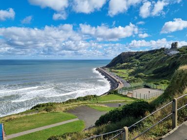 A coastal view with a road along the shore green hills and a fenced sports court at The Seahome in Scarborough