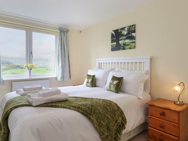 A bedroom with a white bed with green pillows and blanket beside a wooden nightstand with a lamp and a window with curtains and vase at Chase View in Bowland Bridge near Bowness-On-Windermere