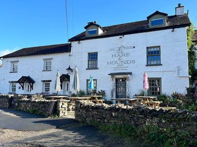 A white stone building with signs for The Hare and Hounds Inn with outdoor wooden picnic tables and closed umbrellas at Bowland Bridge near Bowness-On-Windermere