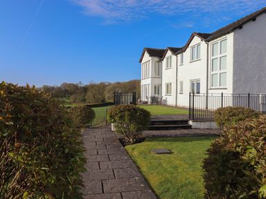 A pathway lined with bushes leading to a white building with multiple windows and outdoor seating at Chase View in Bowland Bridge near Bowness-On-Windermere