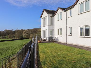 A white house with multiple windows and a metal patio table with chairs on a paved patio beside a green lawn and black metal railing at Chase View in Bowland Bridge near Bowness-On-Windermere