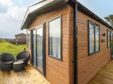 A wooden cabin with black framed windows and doors and black wicker chairs on a wooden deck at The Waves in Little Haven near Broad Haven